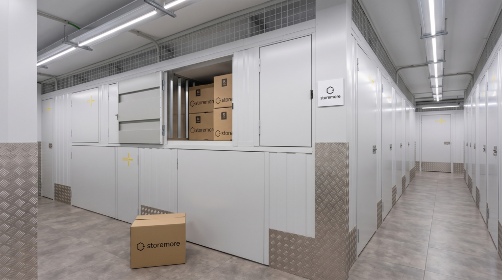 Interior de un trastero storemore con puertas modulares grises numeradas, cajas de cartón apiladas y suelo industrial gris
